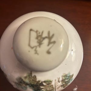 Pu&ograve; includere: Un coperchio in ceramica bianca con un pomello rotondo con un carattere asiatico. Il coperchio presenta un disegno paesaggistico dipinto in verde, blu e marrone. Il coperchio &egrave; probabilmente per un barattolo o un contenitore.