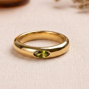May include: A gold ring featuring an olive-green gemstone in a marquise cut. The ring is set against a neutral background, with a lit candle in a glass holder in the background. The ring is a piece of jewellery.