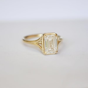 Puede incluir: Un anillo de compromiso de oro con un diamante talla esmeralda. El anillo presenta un engaste detallado y un diseño de caña dividida. El diamante es claro y refleja la luz, sobre un fondo brillante.