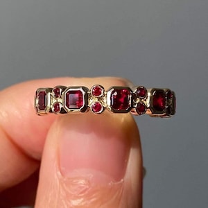 May include: A gold band ring with a row of red ruby gemstones. The rubies are set in a variety of shapes, including square and round.