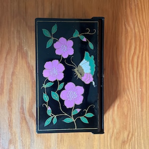May include: A black lacquered box with a floral design featuring pink flowers, green leaves, and a gold butterfly. The box has a hinged lid.