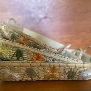 May include: Three woven baskets with floral embroidery in shades of white, green, orange, and gold. The baskets are stacked on top of each other.