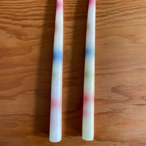 May include: Two white taper candles with multi-colored stripes. The stripes are red, blue, yellow, and green.