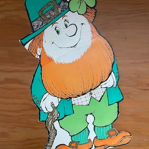 May include: A green leprechaun cutout with a green hat, orange beard, and a shamrock on his hat. He is holding a brown walking stick and is wearing green pants and orange shoes.