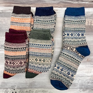 May include: A collection of patterned socks in various colors, including burgundy, navy, and olive green. The socks feature geometric and tribal-inspired designs with a ribbed cuff and heel. The socks are displayed on a wooden surface.