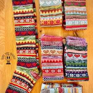 May include: A collection of colorful knit socks with various patterns, including hearts, geometric shapes, and stripes. The socks feature a mix of red, green, yellow, and white designs. The image includes a knee-high sock and several pairs of folded crew socks.