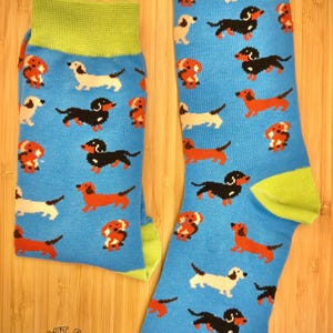 May include: A pair of blue socks featuring a repeating pattern of dachshund dogs in different colours. The socks have green cuffs and heels. The socks are displayed on a wooden surface.