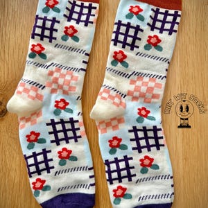 May include: A pair of patterned socks with a light blue and white background. The socks feature a repeating pattern of red flowers, purple grid shapes, and pink and white checkered squares. The cuff is brown, the toe is white, and the heel is purple.