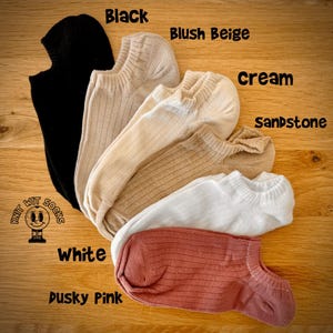 May include: Six pairs of ribbed knit socks in black, blush beige, cream, sandstone, white, and dusky pink. The socks are displayed on a wooden surface. The brand logo "Knit Wit Socks" is also visible.