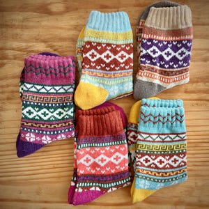 May include: Six pairs of patterned socks in various colours are displayed on a wooden surface. The socks feature geometric designs in shades of purple, red, blue, yellow, and brown. The socks are made of a knitted material.