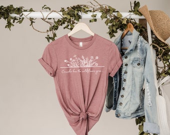 May include: A dusty rose t-shirt with a white graphic of wildflowers and the text "Consider how the wildflowers grow..."
