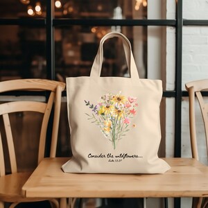 May include: A beige tote bag with a watercolour floral design. The design features yellow, pink, and purple flowers. The text "Consider the wildflowers... Luke 12:27" is printed below the flowers. The bag has two handles.