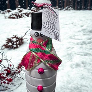 May include: A snowman-shaped bottle with a black top hat, a red and green scarf, and pink button accents. A tag is attached to the top with text. The snowman is set against a snowy winter backdrop with pine cones.