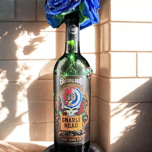 May include: A wine bottle with a label featuring a skull and roses design, topped with a blue rose arrangement. The label reads "Grateful Dead 60th Anniversary Edition" and "Gnarly Head Old Vine Zin". The bottle sits on a dark coaster.
