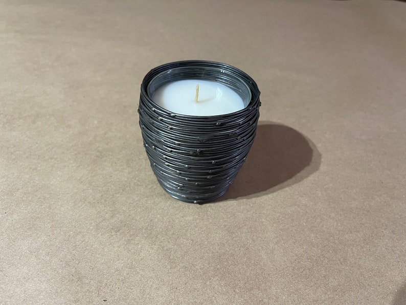 May include: A black candle with a white wax top in a wire container. The wire container has a textured design.