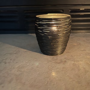 May include: A black metal candle holder with a woven design. The candle holder is empty and has a light shining through the woven design.