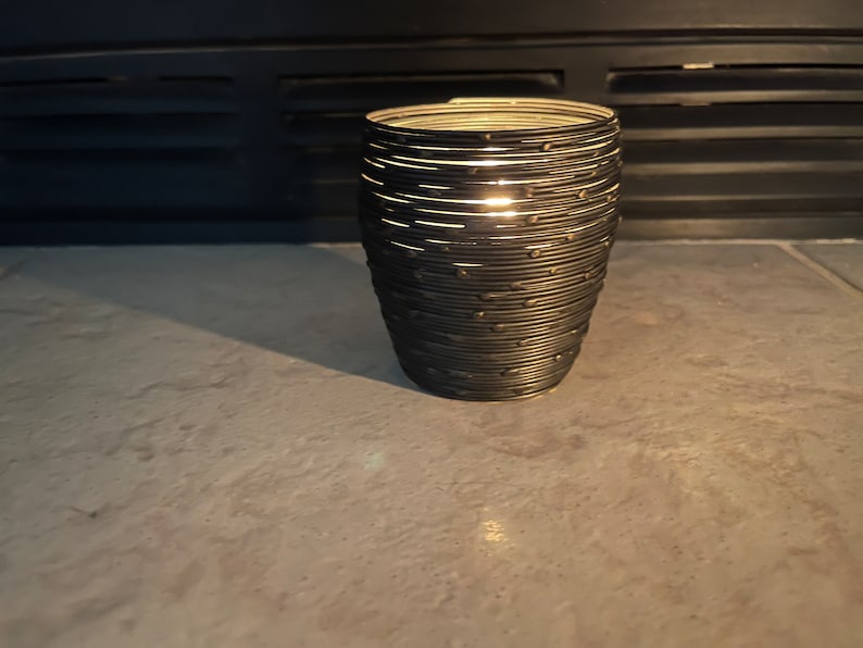 May include: A black metal candle holder with a woven design. The candle holder is empty and has a light shining through the woven design.