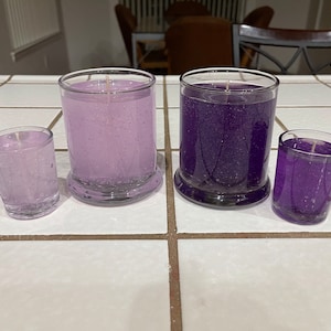 Könnte beinhalten: Vier Glaskerzen in verschiedenen Größen, zwei sind lila und zwei sind hellrosa. Die Kerzen stehen auf einer weißen Fliesenfläche.