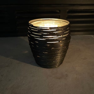 May include: A black metal candle holder with a unique, layered design. The candle holder is lit and casting a warm glow.