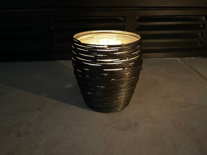 May include: A black metal candle holder with a unique, layered design. The candle holder is lit and casting a warm glow.