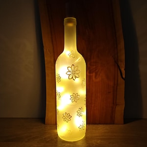 Puede incluir: Una botella de vidrio esmerilado con un diseño floral y luces LED blancas cálidas en su interior. La botella está iluminada y se encuentra sobre una superficie de madera.