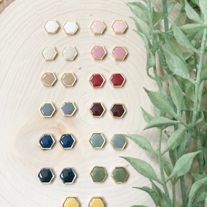 May include: A set of 10 pairs of hexagonal stud earrings in various colors, including white, pink, red, gray, blue, green, and yellow. The earrings are displayed on a wooden surface.