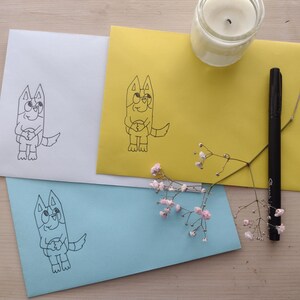 Può includere: Tre buste con un disegno in bianco e nero di un cane dei cartoni animati su ciascuna. Le buste sono bianche, gialle e blu chiaro.