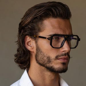 Puede incluir: Gafas cuadradas negras con doble puente y lentes transparentes. Las gafas tienen una montura gruesa y oscura con un acento marrón contrastante en las patillas. La persona lleva una camisa blanca abotonada.