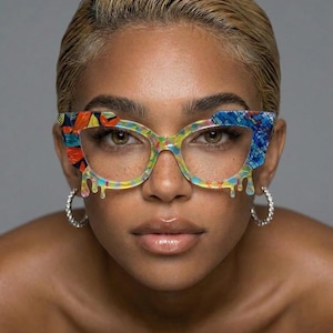 May include: Clear-framed spectacles with colourful patterned accents on the upper corners and a dripping paint design. The glasses have a rainbow colour scheme and are paired with silver hoop earrings. The overall theme is fashion eyewear.