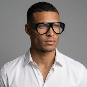 May include: Large, black-framed eyeglasses. The glasses have a bold, rectangular design. The person wearing the glasses is wearing a white shirt. The background is a plain grey.