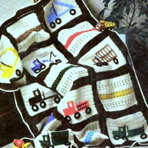 Puede incluir: Una manta de bebé tejida a crochet con fondo blanco y bordes negros. La manta presenta varios vehículos coloridos, incluyendo una excavadora amarilla, un camión de bomberos rojo, un camión azul y un camión de volteo verde.