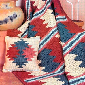 May include: A crocheted throw blanket and pillow with a Southwestern design in shades of red, blue, cream, and pink. The blanket is folded over a wicker chair.