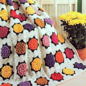 May include: A crocheted blanket with a white background and colorful flower motifs. The blanket is made with a variety of colors, including red, orange, yellow, green, blue, and purple.