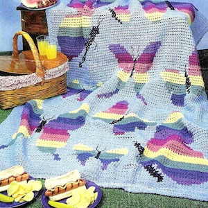May include: A blue crochet blanket with a butterfly design. The blanket is made with multi-coloured yarn and features a rainbow pattern. The blanket is laid out on a green grassy surface with a picnic basket, food, and drinks.