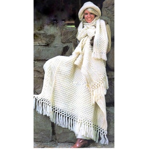 May include: A white crocheted blanket with fringe. The blanket is draped over a person wearing a white crocheted hat and scarf.