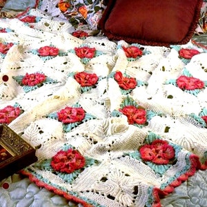May include: A white crocheted throw blanket with a repeating pattern of pink and green flowers. The blanket is folded and draped over a bed.
