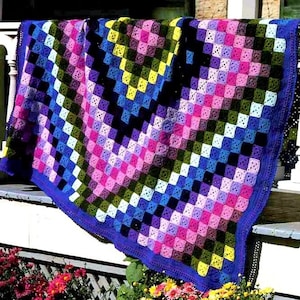 May include: A colourful crocheted blanket with a geometric pattern in shades of pink, purple, blue, green, and black. The blanket has a blue border and is made with a granny square stitch.