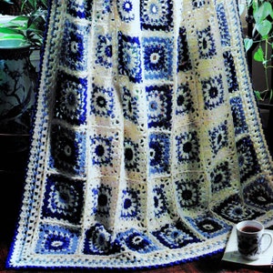 May include: A crocheted blanket with a white background and blue and light blue squares. The blanket is folded and draped over a chair.