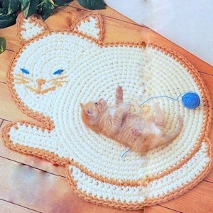 Puede incluir: Una alfombra de crochet con forma de gato con un contorno marrón y un centro blanco. Un pequeño gatito naranja está acostado en la alfombra.