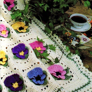 May include: A crocheted blanket featuring a white background and colourful flower designs. The blanket is crafted with a variety of stitches and textures, resulting in a unique and beautiful piece. The flowers are in shades of purple, pink, yellow, and blue.