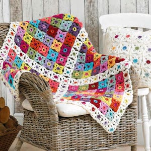 May include: A colourful crocheted throw blanket with a granny square pattern in shades of pink, red, yellow, blue, green, and purple. The blanket is draped over a wicker chair with a white cushion.