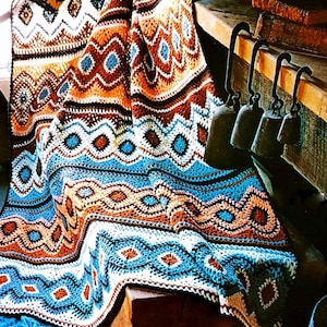 May include: A colorful, crocheted blanket with a geometric pattern. The blanket features a mix of brown, orange, white, and blue hues. The design includes diamond and zigzag shapes, creating a textured and visually appealing textile. The blanket is draped over a wooden surface.