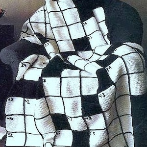 Può includere: Una coperta all'uncinetto in bianco e nero drappeggiata su una sedia. La coperta presenta un motivo a griglia con quadrati numerati, simile a un cruciverba. La coperta è realizzata in filato morbido e ha un aspetto accogliente e fatto a mano.