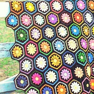 May include: A vibrant, crocheted blanket with a honeycomb design of hexagonal shapes. Each hexagon displays a daisy with bright petals in yellow, orange, pink, blue, and green, set against a black backdrop. The blanket is draped over a white wooden fence.