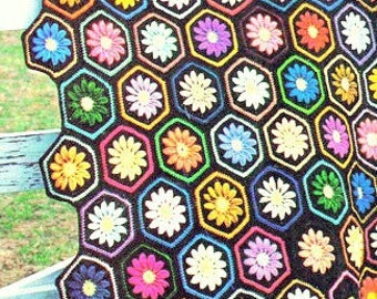 May include: A colorful, crocheted blanket featuring a honeycomb pattern of hexagonal shapes. Each hexagon showcases a daisy with vibrant petals in shades of yellow, orange, pink, blue, and green, set against a black background. The blanket is draped over a white wooden fence.