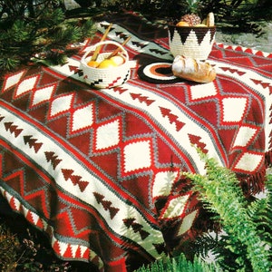 May include: A red, white, and gray crocheted blanket with a geometric pattern. The blanket is laid out on a table with a basket of fruit and a loaf of bread.