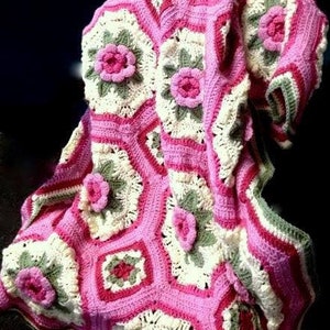 Op de afbeelding: Een gehaakt deken met een roze en wit bloemenpatroon. Het deken is gemaakt van zacht garen en heeft een delicate, vrouwelijke uitstraling.