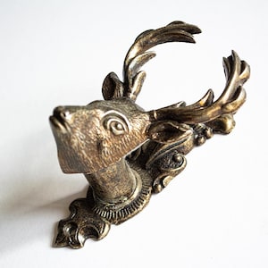 May include: A gold-toned metal deer head wall hook with ornate detailing. The deer's head is facing upwards with its antlers raised.