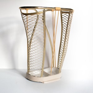 Puede incluir: Un paragüero vintage con estructura metálica dorada y base color crema. El soporte presenta un diseño de malla en forma de diamante y varillas verticales doradas. El asa superior está envuelta con un material de color beige.