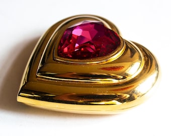 May include: A heart-shaped brooch with a large, faceted, ruby-red gemstone set in a gold-toned metal frame. The brooch has a layered design, with a polished, reflective surface. The background is plain white.